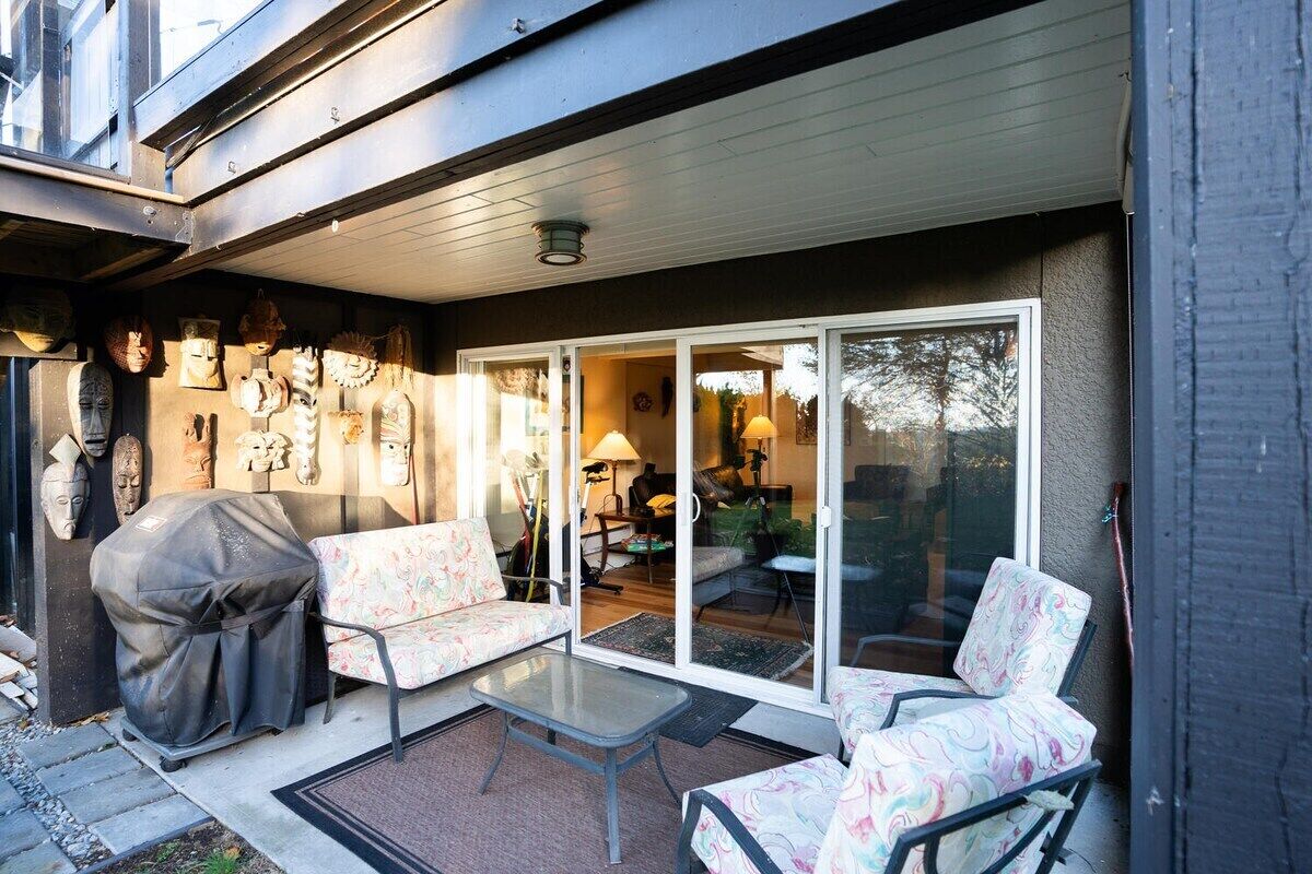 Un patio exterior con una parrilla cubierta, dos sillas estampadas, una mesita y un ventanal corredero que da a un espacio interior.