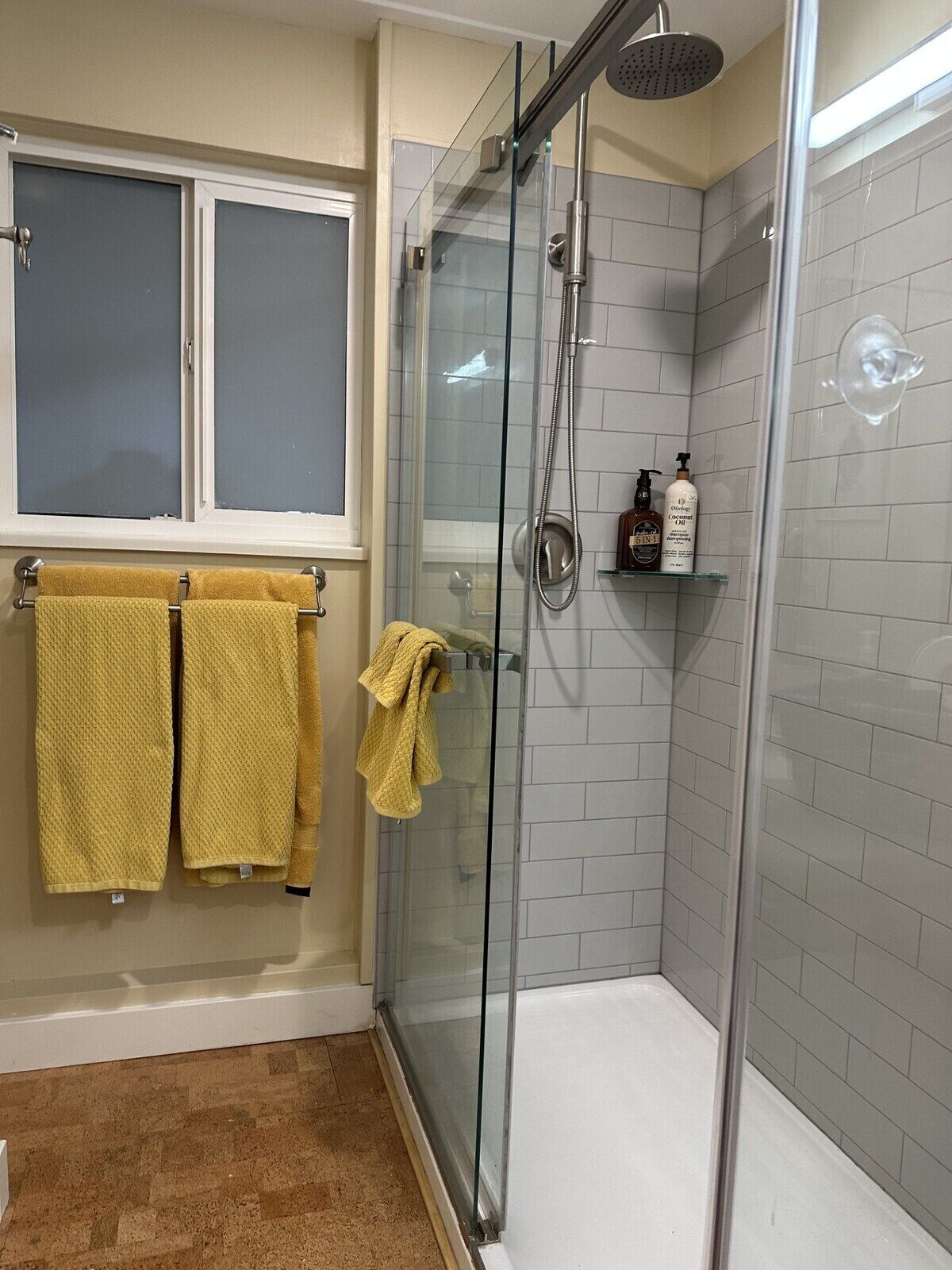 Une salle de bain avec une douche à parois de verre, une fenêtre à carreaux dépolis et un porte-serviettes avec des serviettes jaunes.
