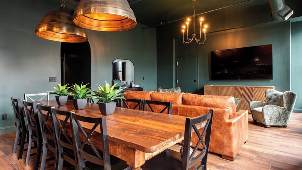 A modern living space with a wooden dining table, leather sofa, and a flat-screen TV mounted on the wall. - Hollyhock Suite 302: 4 Bed/2 Bath (Louisville)