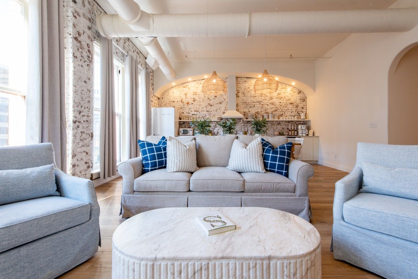 A living room with a sofa, two armchairs, a round coffee table, and a brick wall with a kitchen in the background.