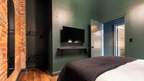 A hotel room with a brick wall, a television, a bed with a quilted cover, and a wooden floor. - Hollyhock Suite 302: 4 Bed/2 Bath (Louisville)