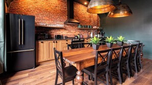 A kitchen with a brick wall, wooden cabinets, a black refrigerator, a wooden dining table with chairs, and hanging pendant lights. - Hollyhock Suite 302: 4 Bed/2 Bath (Louisville)