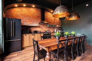 Una cocina con pared de ladrillo, armarios de madera, un refrigerador negro, una mesa de comedor de madera con sillas, y lámparas colgantes.
