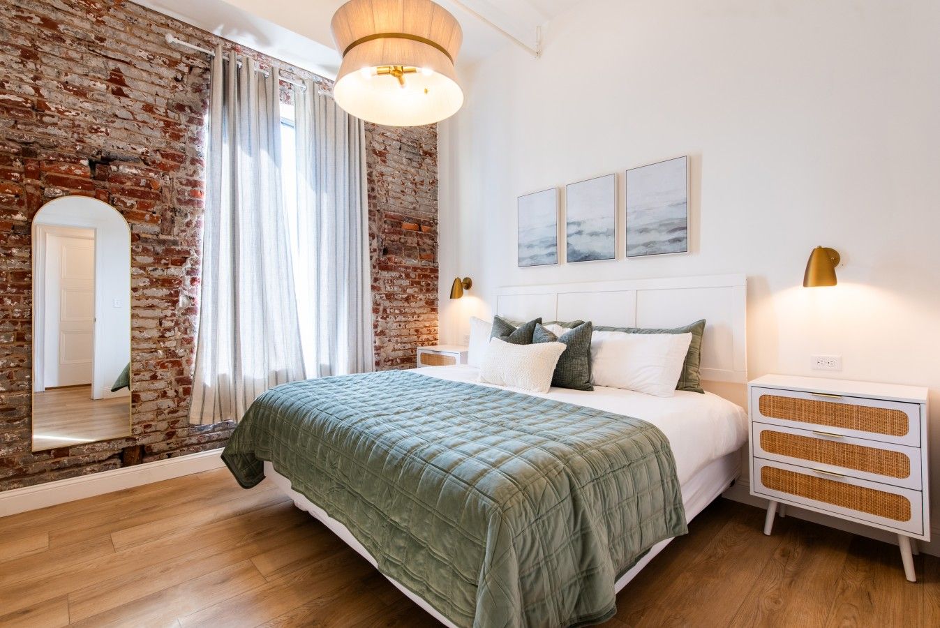 Ein Schlafzimmer mit Backsteinwand, einem Bett mit grüner Bettdecke, einem Nachttisch mit Lampe und einem Spiegel.