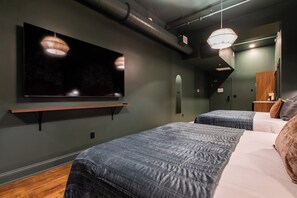 A hotel room with a flat-screen TV mounted on the wall, two beds with blue bedding, a wooden nightstand, and a mirror. - Hollyhock Suite 304: 1 Bed/1 Bath (Louisville)
