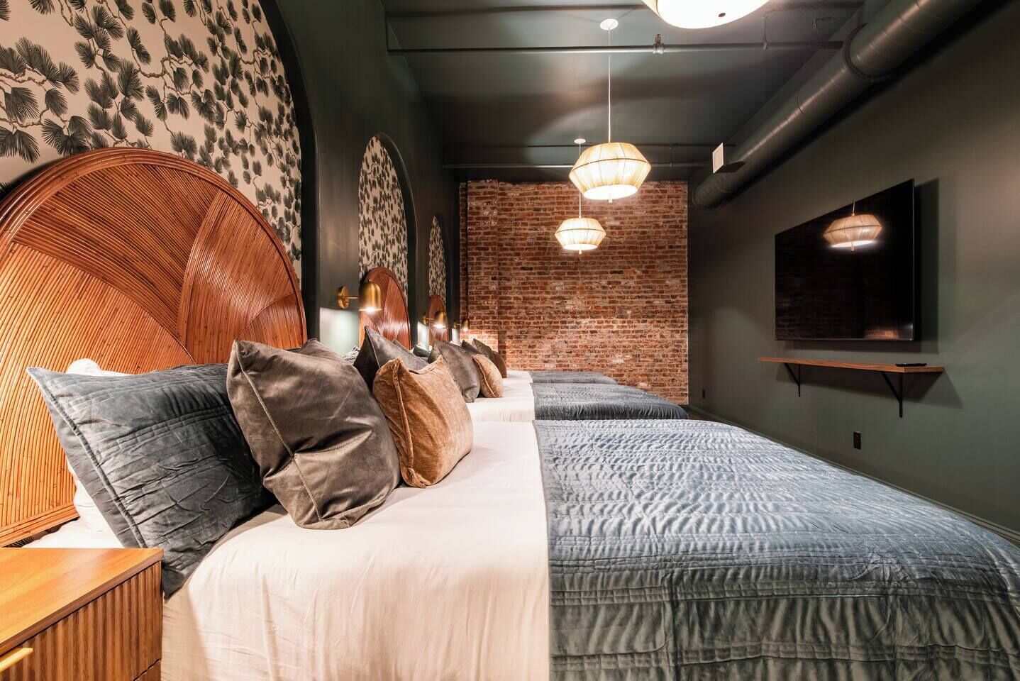 Ein Hotelzimmer mit Backsteinwand, zwei Betten mit grauer Bettwäsche, einem hölzernen Kopfteil, einem an der Wand befestigten Flachbildfernseher und einer Pendelleuchte.