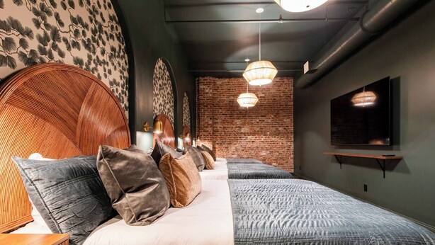 A hotel room with a brick wall, two beds with grey bedding, a wooden headboard, a flat-screen TV mounted on the wall, and a hanging light fixture. - Hollyhock Suite 304: 1 Bed/1 Bath (Louisville)