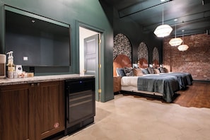A modern kitchen with dark cabinetry and a dishwasher, a bedroom with a large bed and a brick wall, and a dining area with a large table and chairs. - Hollyhock Suite 304: 1 Bed/1 Bath (Louisville)