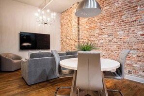 Ein modernes Wohnzimmer mit Backsteinwand, einem runden Esstisch, grauen Sofas und einem Flachbildfernseher.