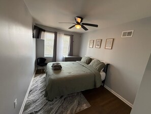 Un dormitorio con cama, escritorio, silla, ventilador de techo y ventana con cortinas.