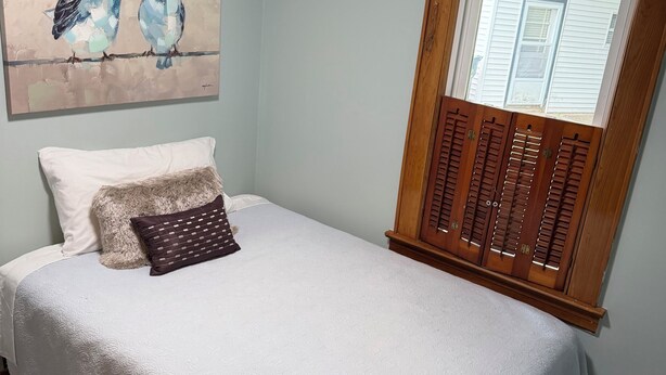 Un dormitorio con cama, un cuadro de pájaros, una ventana con contraventanas y una alfombra estampada.