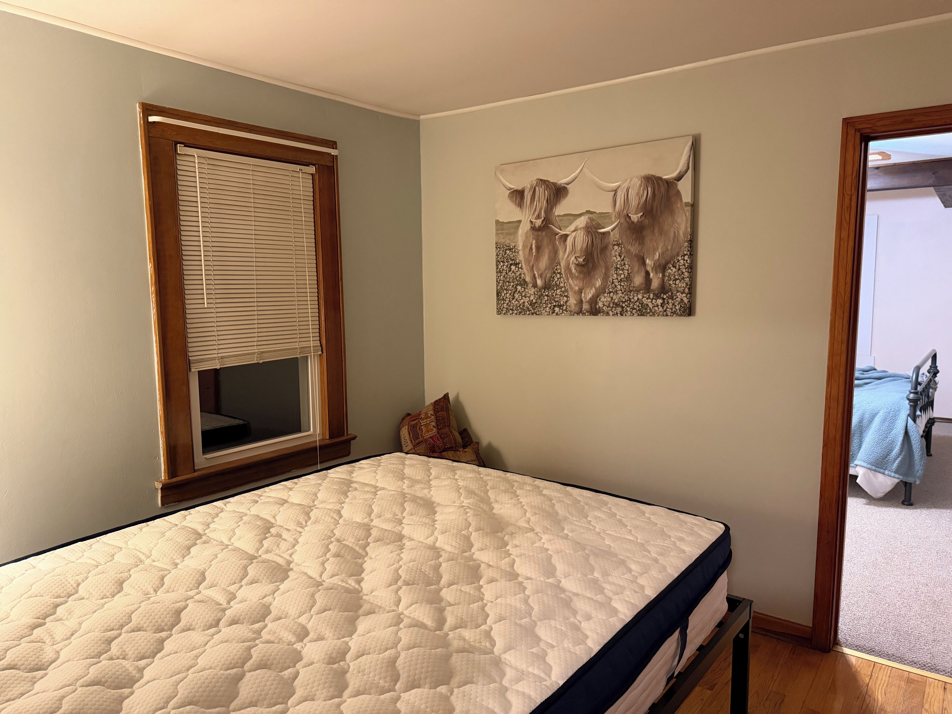 Een slaapkamer met een bed, een raam met zonwering, een schilderij van stieren en een deur die naar een andere kamer leidt.
