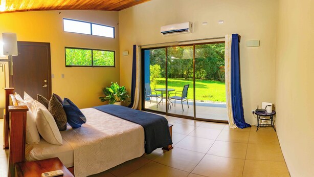 Ein Schlafzimmer mit einem Bett, einem kleinen Tisch, einer Topfpflanze und Blick auf einen Garten durch eine Schiebetür.