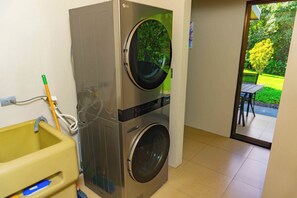 Ein Waschraum mit Waschmaschine und Trockner, einem gelben Waschbecken und Blick auf einen Garten durch eine Glastür.
