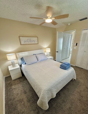A neatly made bed with a quilt, two bedside lamps, a ceiling fan, and a wall-mounted sign. - Coastal Charm, Chic Home Near The Beach! (Port Orange)