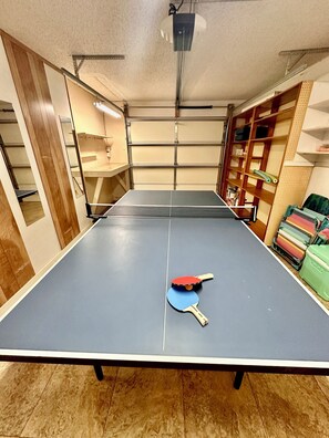 Un tavolo da ping pong con due racchette in una stanza con scaffali in legno e una luce a soffitto.