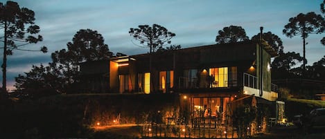 A modern two-story house with a balcony and a lit pool area at dusk.