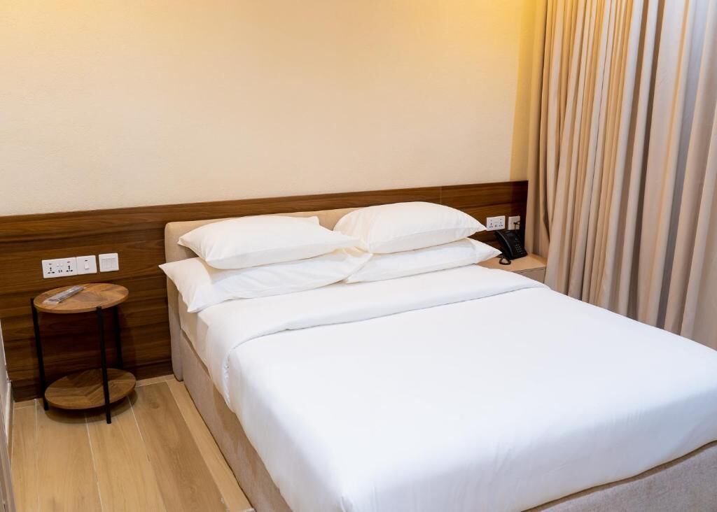 A hotel room with a wooden headboard, a single bed with white linens, a bedside table with a remote control, and a curtain on the window.