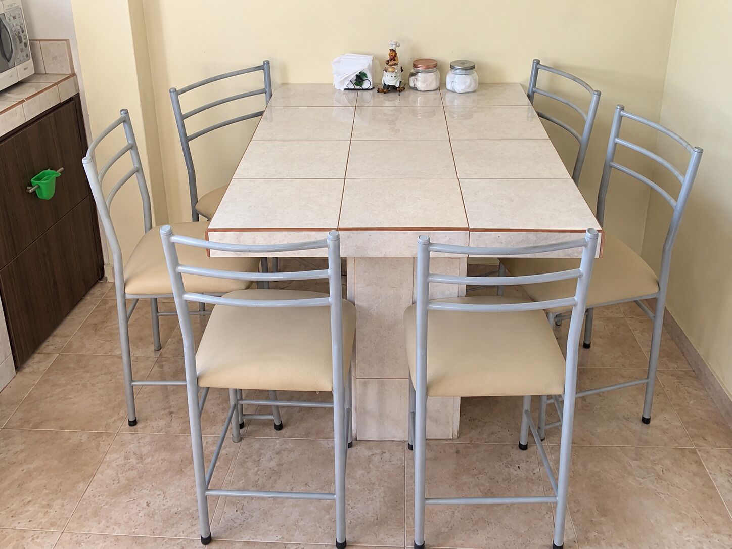 Una mesa de comedor con seis sillas, una zona de cocina con lavadora y una cesta verde en el suelo.