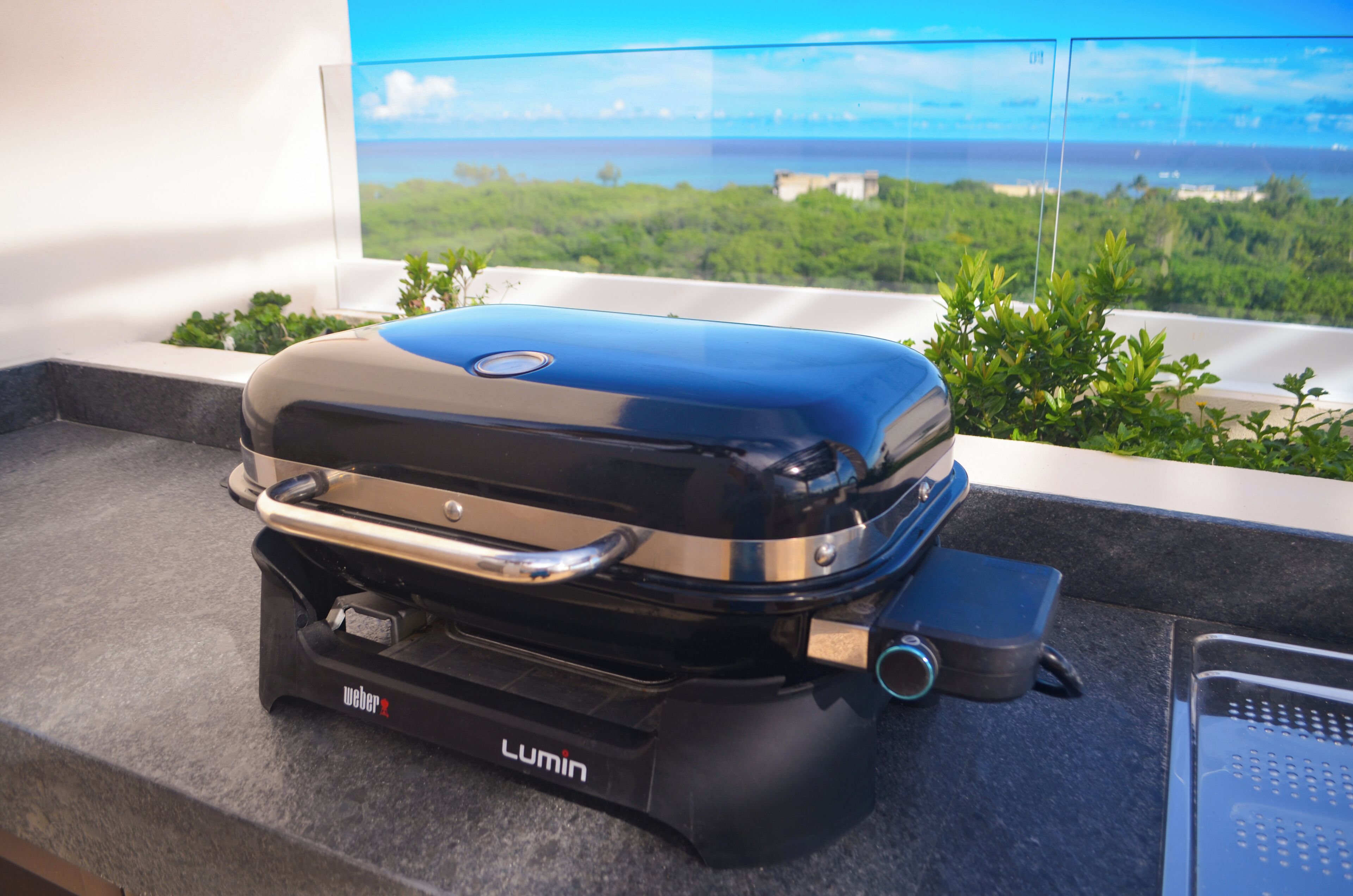 Una parrilla Weber Lumin en un balcón con vista al océano y follaje.