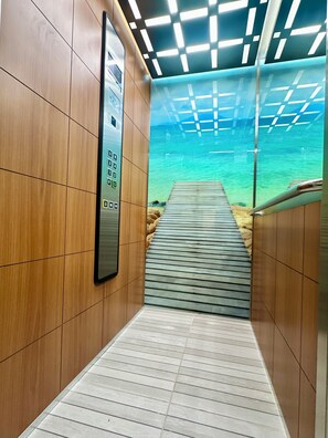 Un pasillo con piso de madera, un panel de ascensor digital y vista a una terraza de madera que da a un cuerpo de agua.
