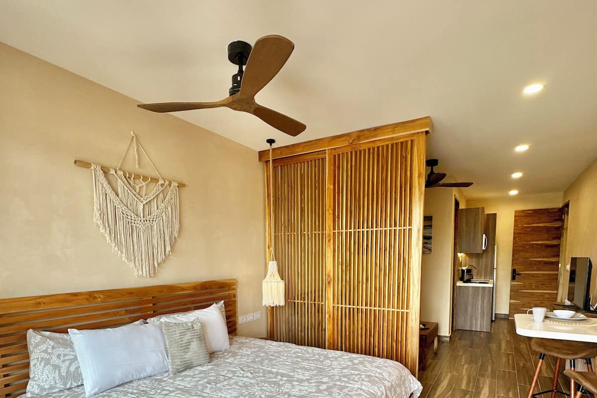 Een ruime slaapkamer met een groot bed, een plafondventilator, een macramé wanddecoratie en uitzicht op het keukenvertrek.