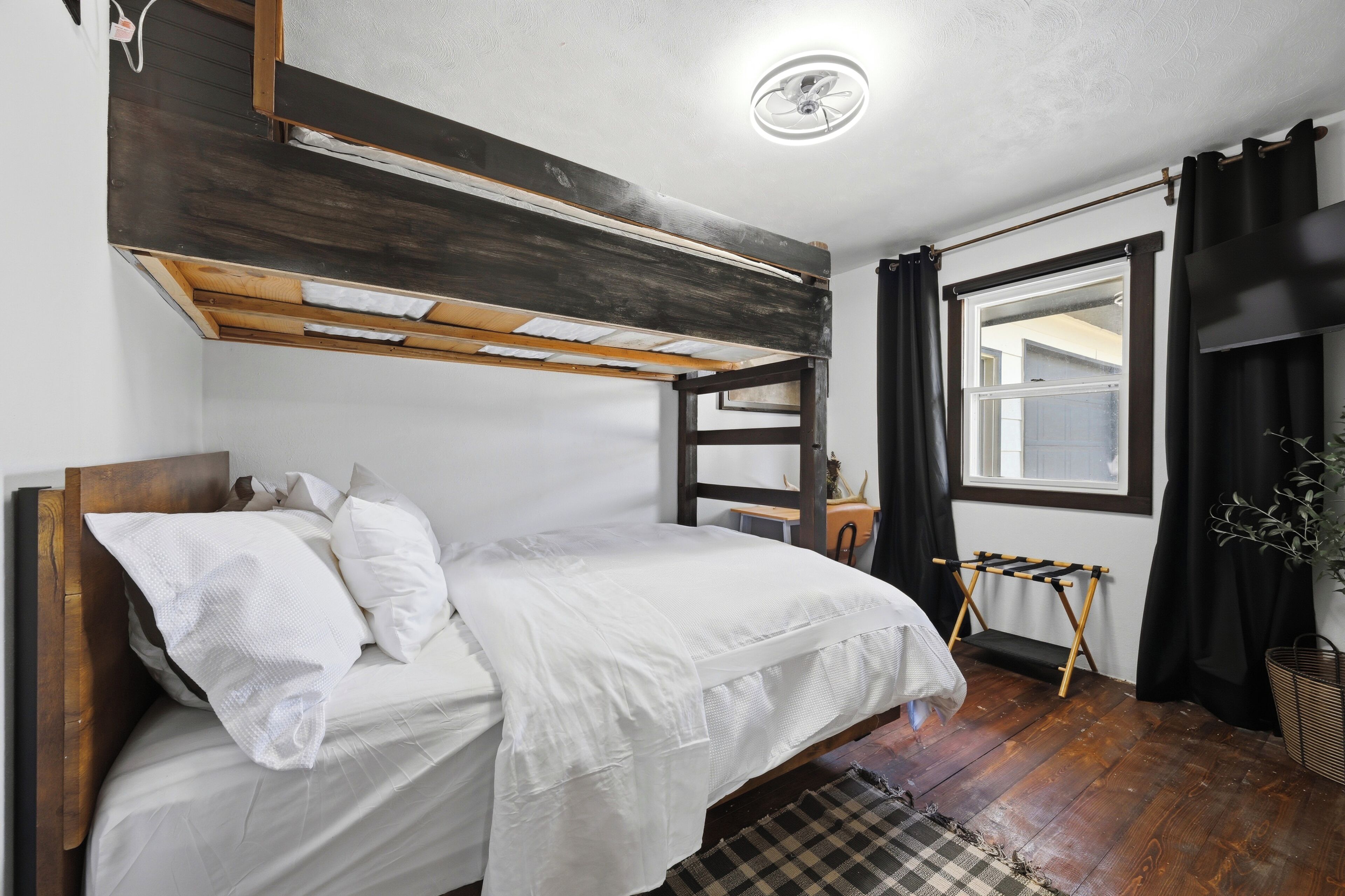 A bunk bed with a wooden headboard, white bedding, a small wooden stool, a window with curtains, and a plant.