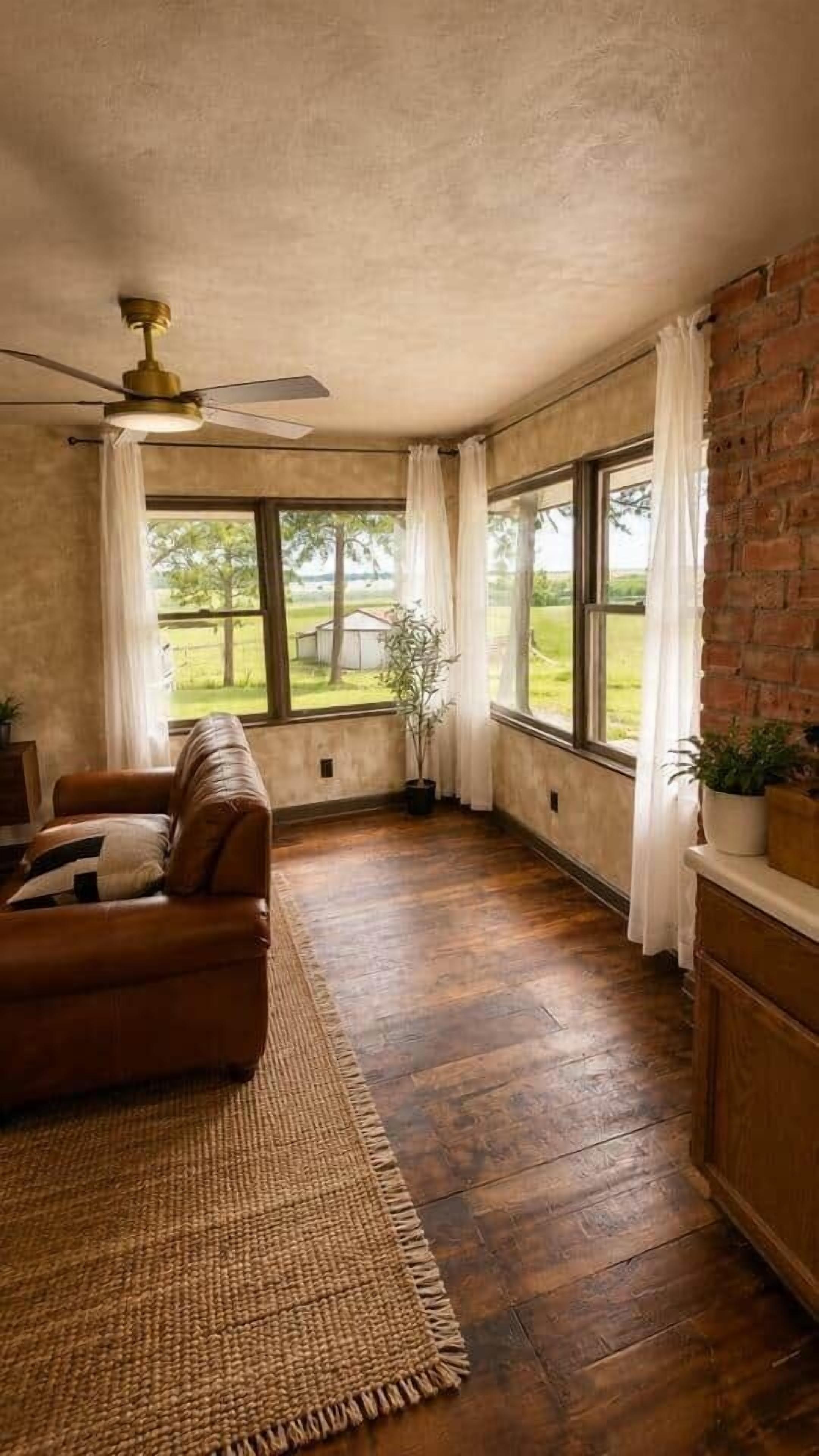 A room with a wooden floor, a brown leather sofa, a ceiling fan, and a brick wall.