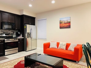 A modern kitchen with dark cabinets, a refrigerator, and a microwave. There is a red sofa with white cushions and a painting of flowers on the wall. - Brand new guest suite in beautiful locality (Mountain House)
