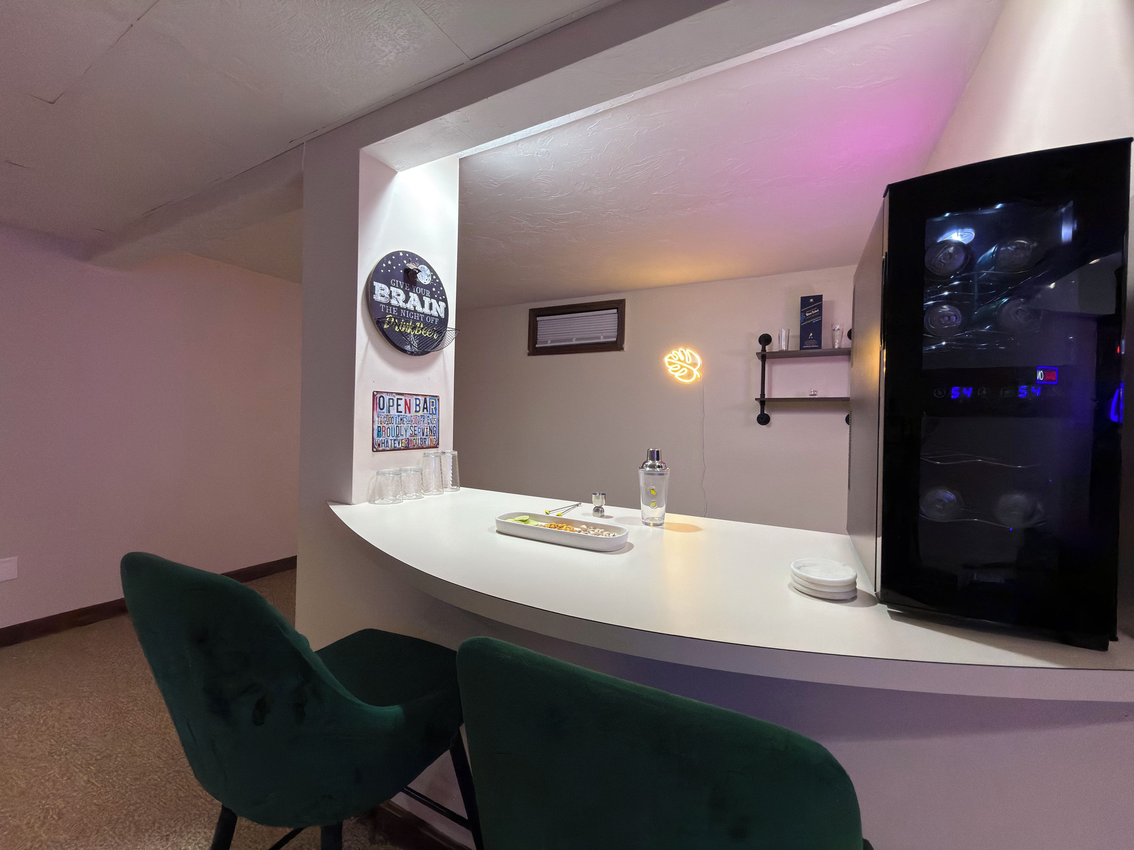 A bar area with a counter, two green chairs, a refrigerator, and a neon sign on the wall.