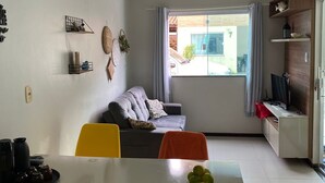A modern living room with a sofa, a dining table with chairs, a TV on a stand, and a window with curtains. - Casa 40, Condomínio Lençóis Ville Residence (Barreirinhas)