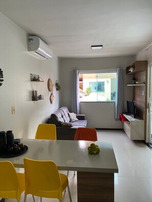 Ein modernes Wohnzimmer mit einer Couch, einem Esstisch mit Stühlen, einem Fernseher auf einem Schrank und einem Fenster mit Vorhängen.