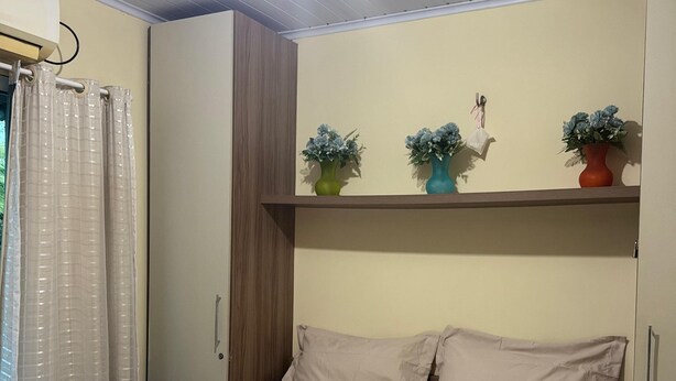 A bedroom with a bed, a shelf with potted plants, and a window with curtains. - Casa 40, Condomínio Lençóis Ville Residence (Barreirinhas)