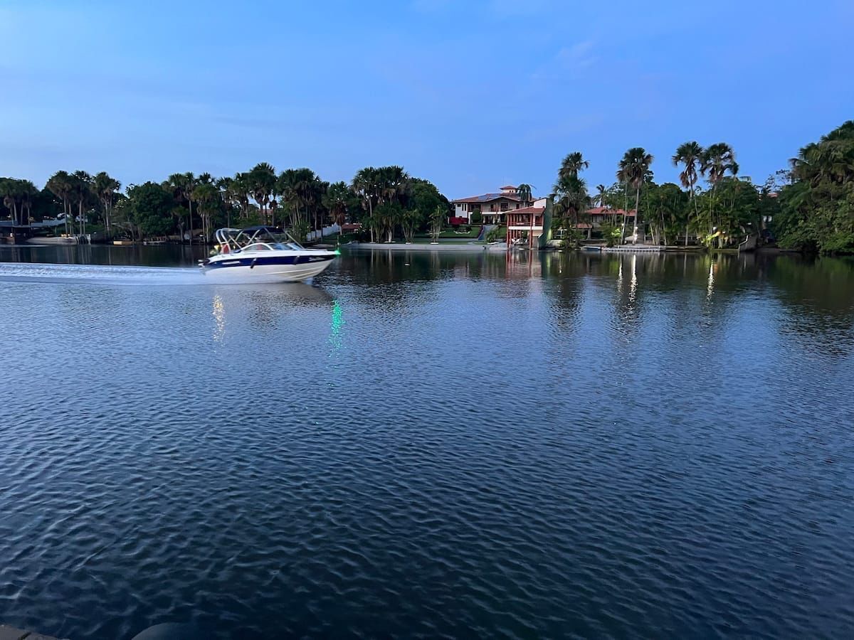 Um lago tranquilo com um barco em movimento, rodeado por árvores e edifícios ao fundo.