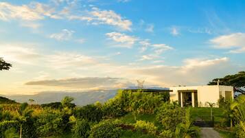 A modern building with a pathway leading to a gated entrance, surrounded by lush greenery and a clear sky.