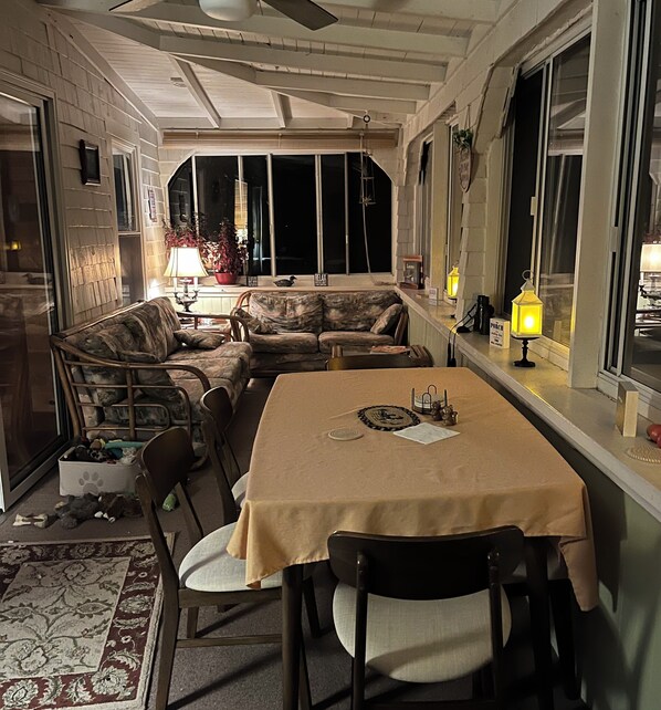 A dining area with a table, chairs, and a sofa. There are potted plants and a window with a view.
