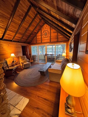 A cozy cabin interior with wooden walls, a stone fireplace, a large window, and a comfortable seating area.