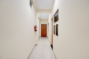 A narrow hallway with a wooden door at the end, white walls, and a fire extinguisher mounted on the wall. - Ollection O Sector 95B Gurgaon (Gurugram)
