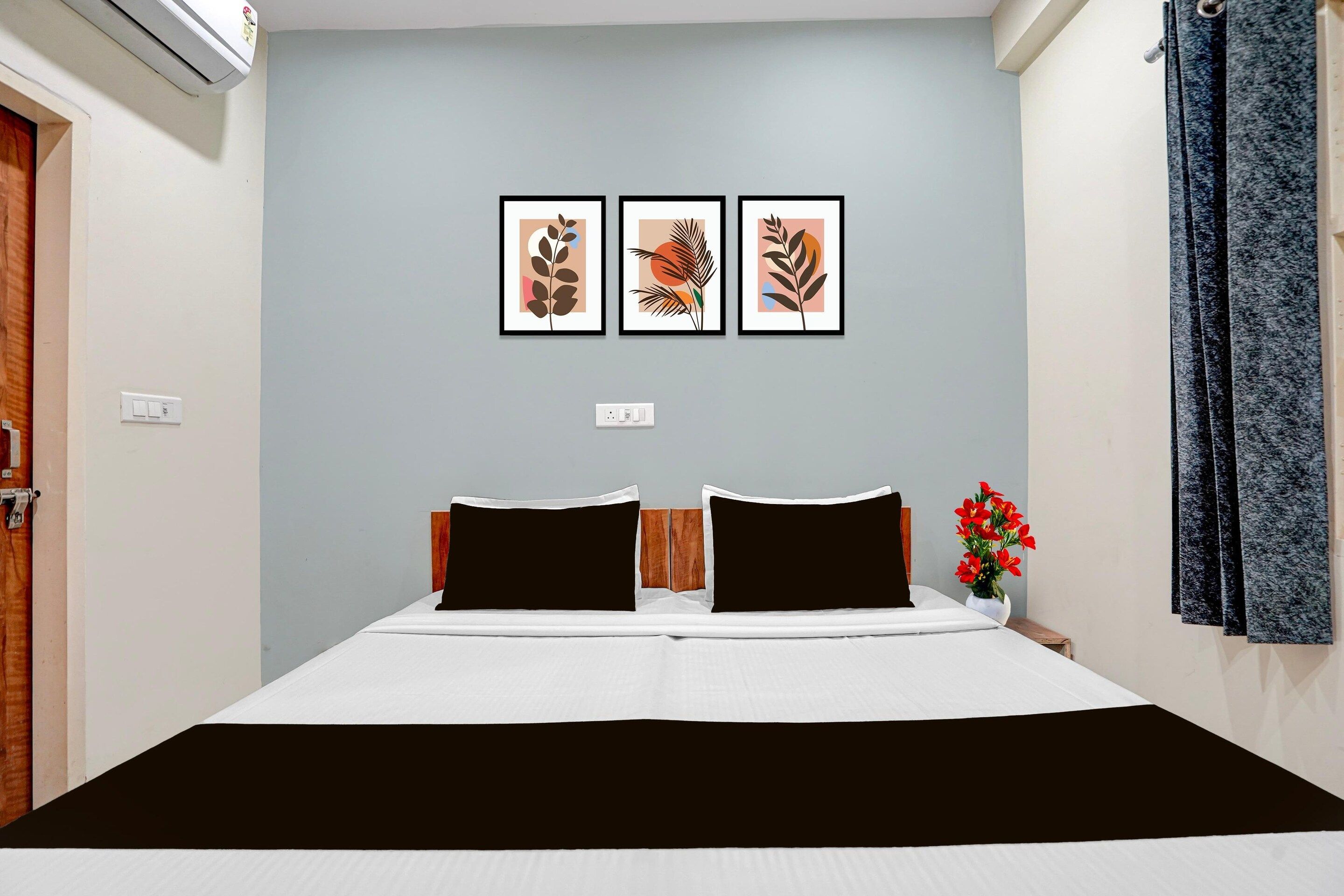 A bedroom with a bed, two pillows, a nightstand with a vase of flowers, and three framed artworks on the wall.