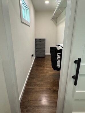Un couloir étroit au plancher en bois, une porte blanche sur la droite et un sac à linge suspendu au mur.
