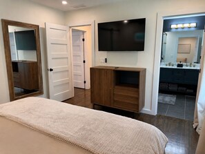 Ein Schlafzimmer mit einem Bett, einem an der Wand befestigten Fernseher, einem Holzschrank und einem durch eine offene Tür einsehbaren Badezimmer.