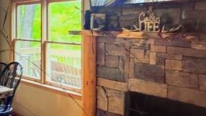 A room with a stone fireplace, a window with a view of greenery, and a chair. - Cabin 38 miles from Beach Mountain ski resort 5 miles from the lake in winery (Butler)