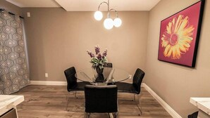 A dining area with a glass table, black chairs, a vase with flowers, a pendant light, and a large flower painting on the wall. - Carlton Suite 1b/1b near UNLV (Las Vegas)