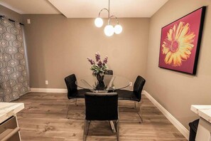 A dining area with a glass table, black chairs, a vase with flowers, a pendant light, and a large flower painting on the wall. - Carlton Suite 1b/1b near UNLV (Las Vegas)