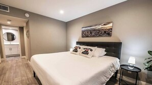 A modern bedroom with a large bed, a bedside table, a round mirror, and a cityscape painting on the wall. - Carlton Suite 1b/1b near UNLV (Las Vegas)