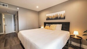 Habitación de hotel con una cama grande, una mesita de noche con lámpara, un cuadro en la pared y una puerta que da a otra habitación.