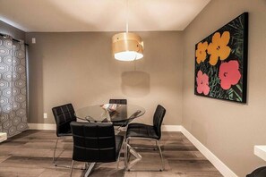 Un comedor con una mesa redonda de cristal, cuatro sillas negras, una lámpara colgante y un cuadro floral en la pared.