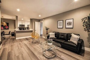 A modern living room with a black leather sofa, a glass coffee table, and a yellow chair. There is a kitchen in the background with white cabinets and a microwave. - Vermont Suite 1b/1b near UNLV (Las Vegas)
