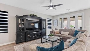 A living room with a large flat-screen TV, a dark entertainment unit, a beige sofa with blue cushions, and a ceiling fan. - Skye Canyon 3 bedrooms + Loft (Las Vegas)
