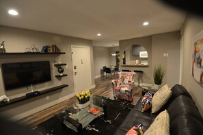 A modern living room with a flat-screen TV, a glass coffee table, a black leather sofa, and a patterned armchair. - Crawford suite 1b/1b near UNLV (Las Vegas)